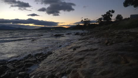 Wide-slow-motion-footage-of-waves-rolling-over-rocks-on-foreshore-while-sun-sets-in-cloudy-sky-during-dusk