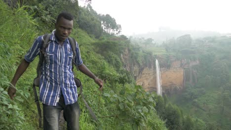 Afrikanischer-Mann,-Der-Auf-Einem-Kleinen-Schlammigen-Pfad-In-Einem-Tropischen-Dschungel-Spaziert,-Mit-Einem-Großen-Wasserfall,-Der-Von-Einer-Klippe-Im-Hintergrund-Fällt
