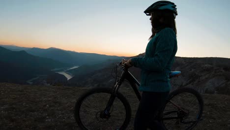 Girl-pushing-her-bike-on-a-mountain-hill-at-sunset-with-beautiful-canyon-lake-in-the-background
