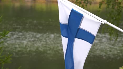 The-flag-of-Finland-blowing-in-the-wind-at-countryside-cabin-by-the-lake,-slow-motion-tilting-up