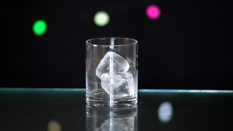 Riesige-Eiswürfel-Fallen-In-Ein-Schnapsglas,-Das-Auf-Einer-Glasoberfläche-Mit-Schwarzem-Hintergrund-Und-Lichterketten-Sitzt