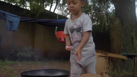 A-young-mixed-raced-boy-throws-a-sausage-on-a-frying-pan-over-a-camp-fire-in-his-backyard