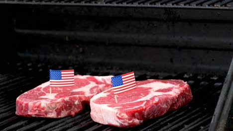 Zwei-Saftige-Rib-Eye-Steaks-Sitzen-Auf-Dem-Grill-Und-Kochen-Eine-Hand-Kommt-Herein-Und-Steckt-Mit-Einem-Zahnstocher-Eine-Winzige-Amerikanische-Flagge-In-Eines-Von-Ihnen
