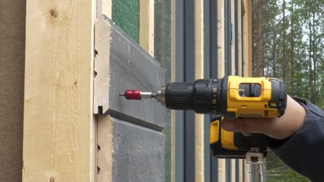 Male-Hand-Holding-Electric-Screwdriver-And-Screwing-interlocking-wooden-timber-planks-onto-house-facade,-tongue-and-groove