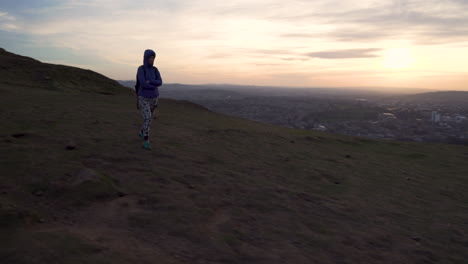 Circling-parallax-shot-of-girl-walking-on-top-of-the-Arthurs-seat-mountain-on-the-hiking-trail-in-evening,-dusk,-blue-hour-with-cityscape-of-Edinburgh-in-the-background