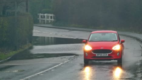 Rotes-Auto-Spritzt-Welle-Auf-Stürmischer,-Von-Sturzfluten-überschwemmter-Straßenecke-In-Großbritannien