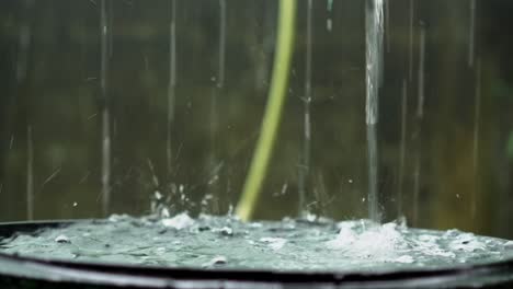 Rain-droplets-falling-and-filling-up-rainwater-harvesting-barrel,-closeup