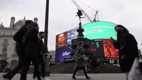 A-woman-wearing-a-surgical-face-mask-walks-past-the-Eros-Statue-and-the-neon-advertisement-billboard-sign-at-Piccadilly-Circus