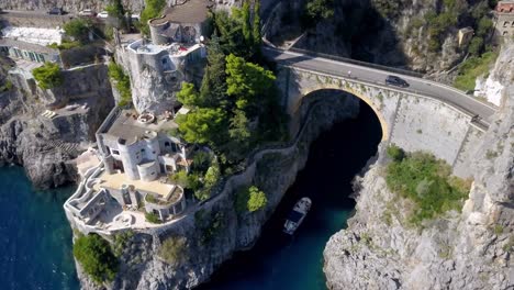 Fiordo-di-Furore-arch-bridge-in-the-Italian-Almalfi-coast-with-car-traffic-above-and-boat-entering-below,-Aerial-lowering-approach-shot
