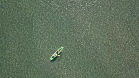 two-people-in-sit-on-top-kayak-paddle-in-shallow-green-water,-aerial