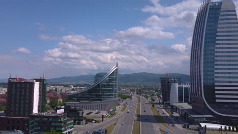 Drone-flying-forward-passing-modern-business-buildings-above-traffic-road