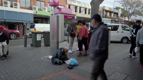 An-old-Asian-beggar-or-homeless-is-eating-while-passerby-people-are-staring-and-preparing-to-give-money-at-Cabramatta,-Sydney,-Australia