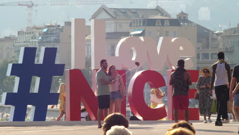 Hübsches-Paar,-Das-Mit-Einem-Einbeinstativ-Am-Schild-„I-Love-Nice-Background“-In-Nizza,-Frankreich,-Ein-Foto-Macht