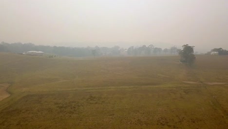 Thick-smoke-in-the-air-engulfs-ranch-area-with-grassy-plains-in-fire-danger-zone-from-eucalyptus-forest-blaze,-Aerial-drone-flyover-reveal-shot