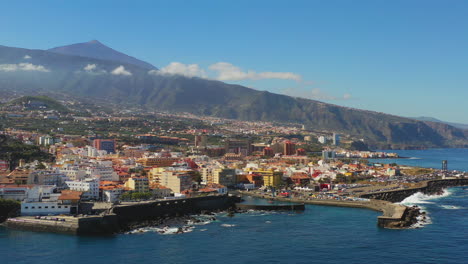 Scenic-view-of-the-vibrant-city-and-harbor-of-Puerto-de-la-Cruz,-Canary-Islands,-Spain,-on-the-coast-of-the-Atlantic-ocean,-Pico-de-Teide-mountain-in-the-background,-blue-sea-and-sky,-aerial-shot-4K