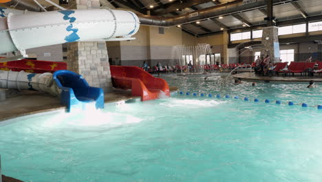 Panning-footage-of-an-indoor-swimming-pool,-fountain,-and-kids-playing,-with-two-waterslides