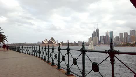 Walking-shot-under-Harbour-Bridge-with-Opera-House-and-downtown-buildings-in-the-distance-on-an-overcast-day,-Stabilized-pan-left-shot