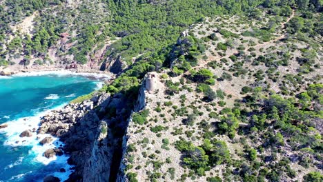 Basset-Cove-in-Mallorca-Spain-with-tower-overlooking-the-blue-cove-below,-Aerial-dolly-out-reveal-shot
