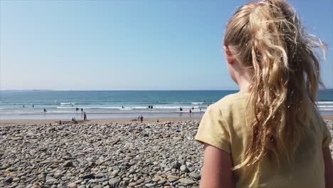 Seven-year-old-blond-girl-in-yellow-t-shirt-climbing-pebble-bank-to-reveal-busy-beach,-sea---horizon-on-clear-summers-day