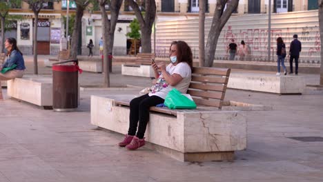 Lady-with-face-mask-using-cellphone,-sitting-on-Merced-square