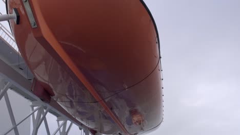 Orange-lifeboat-hanging-on-offshore-vessel,-viewed-from-the-bottom-against-grey-cloudy-sky