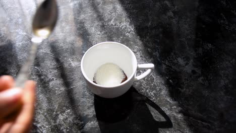 Zucker-In-Eine-Weiße-Tasse-Auf-Einem-Schwarz-Marmorierten-Tisch-Löffeln,-Während-Sonnenstrahlen-über-Die-Szene-Fallen