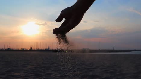 Hand-Lässt-Etwas-Sand-Fallen,-Mit-Sonnenuntergang-Im-Hintergrund-Am-Strand