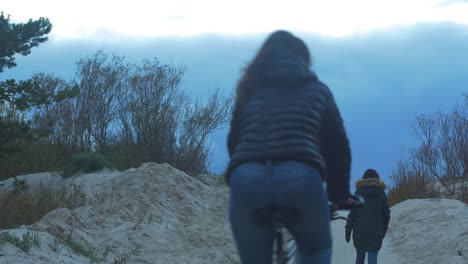 Chica-Morena-Y-Dos-Personas-Con-Bicicletas-Cruzando-Dunas-Junto-Al-Mar