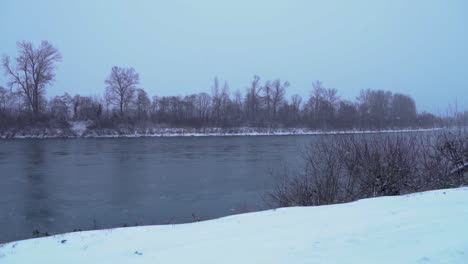 Snow-falling-in-storm-by-river,-Slow-Motion,-Pan-to-Left