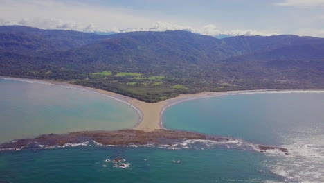 Aerial-shot-pulling-away-from-the-whale-tail-shaped-rocky-point-of-Punta-Uvita-surrounded-by-bright-blue-ocean-waves-and-mountains-in-the-distance-in-south-Puntarenas,-Costa-Rica