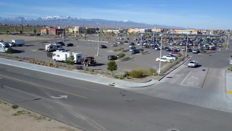 Aerial-footage-of-a-Walmart-parking-lot-in-Adelanto,-California-showing-parked-recreational-vehicles-with-cars-moving,-people-walking,-businesses,-and-snow-capped-Angeles-Mountains-in-the-background