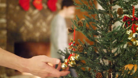 Eine-Person-Hängt-Ein-Jinglebell-Ornament-An-Einen-Weihnachtsbaum-Mit-Einem-Feuer-Und-Einem-Strumpf-über-Dem-Kamin-Für-Die-Feiertage