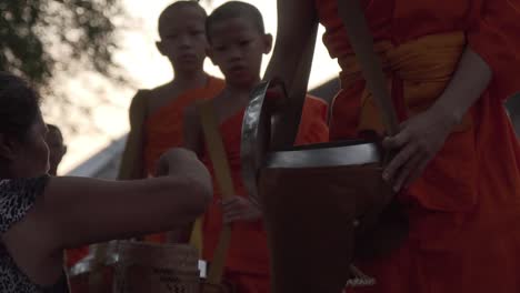 Jóvenes-Monjes-Budistas-Vestidos-Con-Una-Túnica-Naranja-Tradicional-Por-La-Mañana-En-La-Calle-Y-Una-Mujer-Laosiana-Asiática-Alimentan-A-Los-Monjes-Con-Arroz-Dentro-De-Una-Cesta-De-Bambú