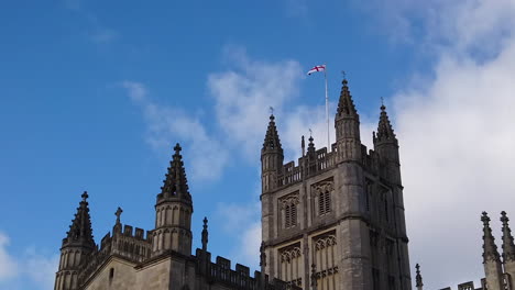 Super-Slow-Motion-Shot-of-Saint-George’s-Cross-Flapping-in-the-Wind-on-top-of-the-Tower-of-Bath-Abbey-in-Somerset,-England-on-Sunny-Summer’s-Day-with-Blue-Sky