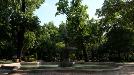 People-walking-around-beautiful-fountain-in-a-park-in-Plovdiv,-European-capital-of-culture