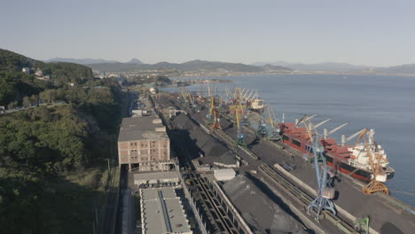 Aerial-shot-of-a-coal-port-with-moored-general-cargo-bulk-carrier-ships-and-a-bay-with-green-hill-side,-on-a-bright-sunny-day