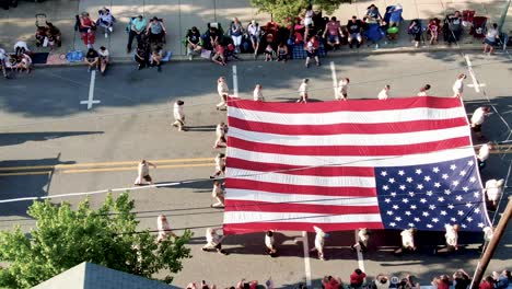 Aerial-truck-shot-of-Boy-Scouts-of-America-with-flag-in-parade,-Cub-Scouts-follow,-crowds-cheering-along-street-on-sidewalk