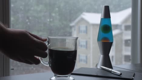 hand-places-glass-mug-of-coffee-down-snowing-outside-home-office-apartment-slow-motion