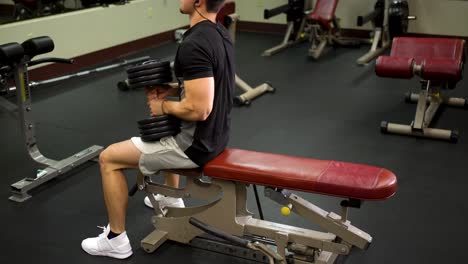 Side-shot-of-teen-bodybuilder-adjusting-himself-on-a-bench-and-lifting-a-heavy-dumbbell-over-his-head
