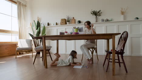 Wide-shot-of-a-mother-making-a-videocall-while-her-daughter-is-distracted-under-the-table-drawing