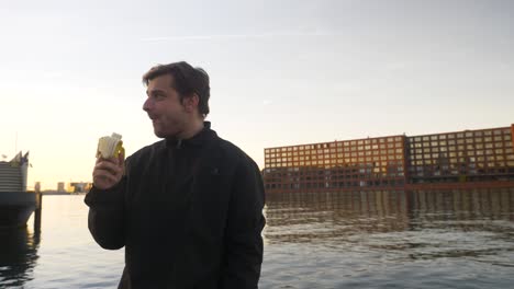 Wide-shot-of-a-young-man-eating-a-banana-and-rejoicing-on-a-pier-during-sunset