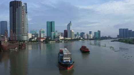 drone-shot-of-A-cargo-ship-and-smaller-vessel-are-transporting-shipping-containers-along-the-Saigon-river-with-the-financial-and-city-center-in-full-view