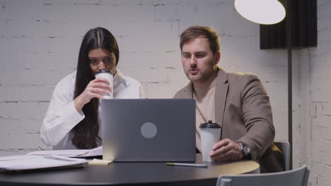 Man-and-woman-having-a-business-meeting-sitting-at-desk-in-boardroom.-They-analyzing-data-together-on-laptop-computer-while-drinking-takeaway-coffee.