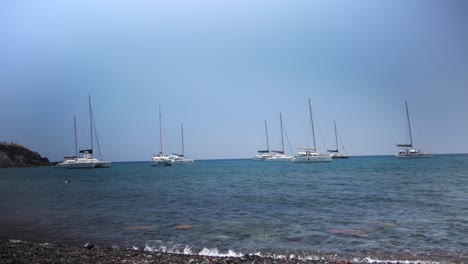Handheld-Wide-Shot-of-Eight-Sail-Boat-Yachts-Gathered-By-Red-Beach-in-Santorini-Greece-and-a-Guy-Swimming