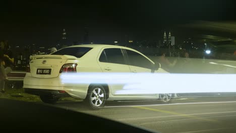 White-Car-Parked-On-The-Roadside-In-Kuala-Lumpur,-Malaysia-With-Cars-Passing-By-On-The-Road-At-Night---timelapse