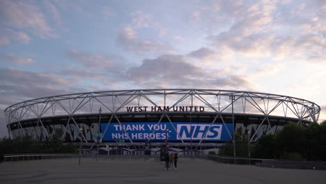 A-time-lapse-of-clouds-passing-over-the-Olympic-Park-Queen-Elizabeth-Stadium-at-sunset,-where-a-giant-billboard-praises-NHS-healthcare-workers-and-warns-the-public-to-maintain-social-distancing