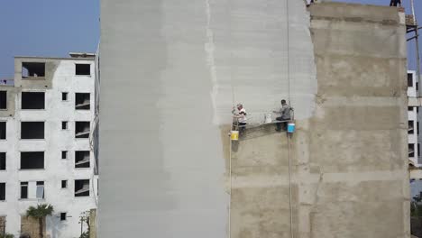 Construction-workers-hanging-on-the-side-of-a-building-applying-concrete-plaster-finish,-Aerial-orbit-around-shot