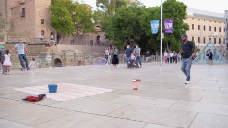 Man-With-Mask-Entertaining-Kids-With-A-Huge-Bubble-Wand-In-Front-Of-Roman-Theatre-In-Malaga,-Spain---wide-shot