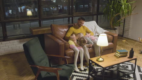 Happy-Caucasian-father-sitting-on-sofa-with-his-two-sleepy-little-girls-who-leaning-their-heads-on-his-lap