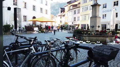 Flowers-display-with-people,-parked-bicycle,-building-and-monument-in-town-of-Feldkirch-Vorarlberg-Austria-during-covid-19